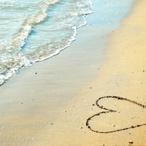 A heart drawn in the sand with waves approaching on a sunny beach day.