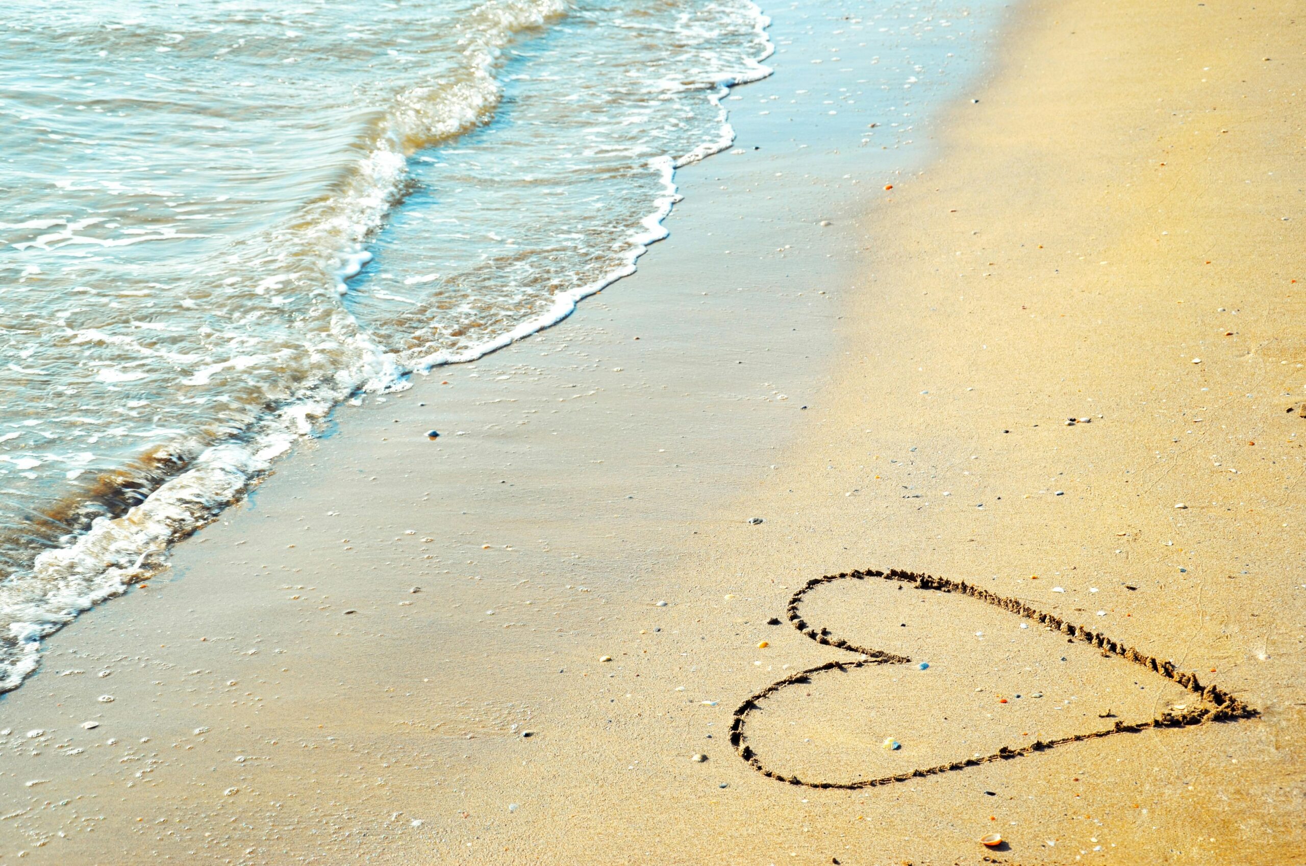 A heart drawn in the sand with waves approaching on a sunny beach day.